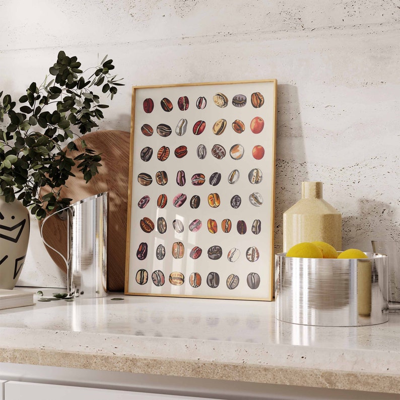 May include: A framed print with a white background and a grid of colorful illustrations of coffee beans. The print is on a white countertop with a silver pitcher and a silver bowl with lemons.