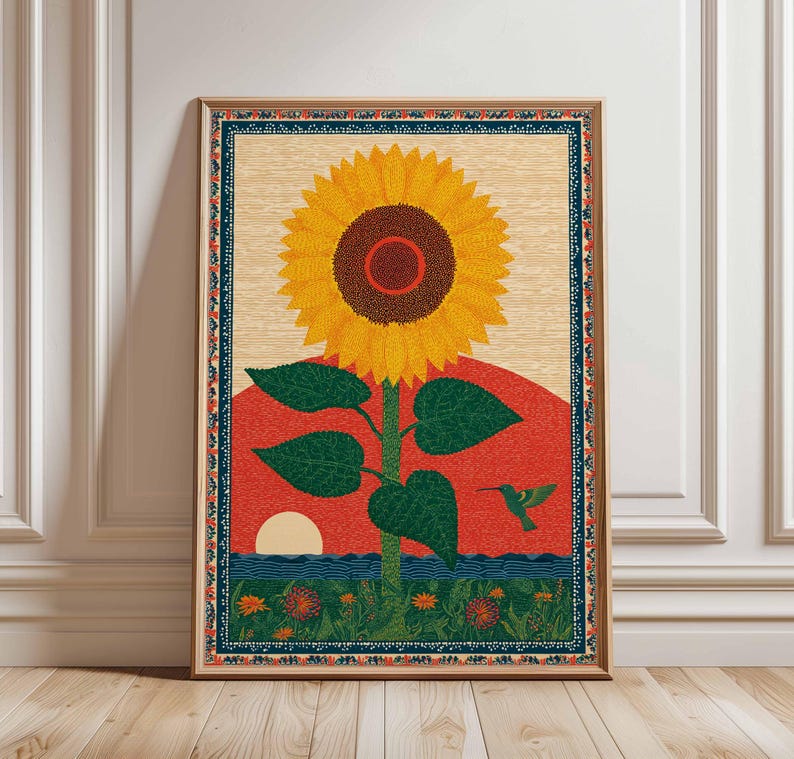 Op de afbeelding: Een ingelijste kunstdruk met een grote zonnebloem met gele bloemblaadjes en een bruin hart. Het kunstwerk bevat een rode zonsondergang, blauw water en een groene kolibrie. De print heeft een decoratieve rand en een lichte houten achtergrond.