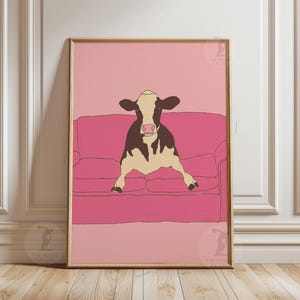 May include: Framed art print showing a cow relaxing on a pink sofa. The cow is illustrated in black and white, set against a pink backdrop and sofa. The artwork has a minimalist, modern style, ideal for home decor.