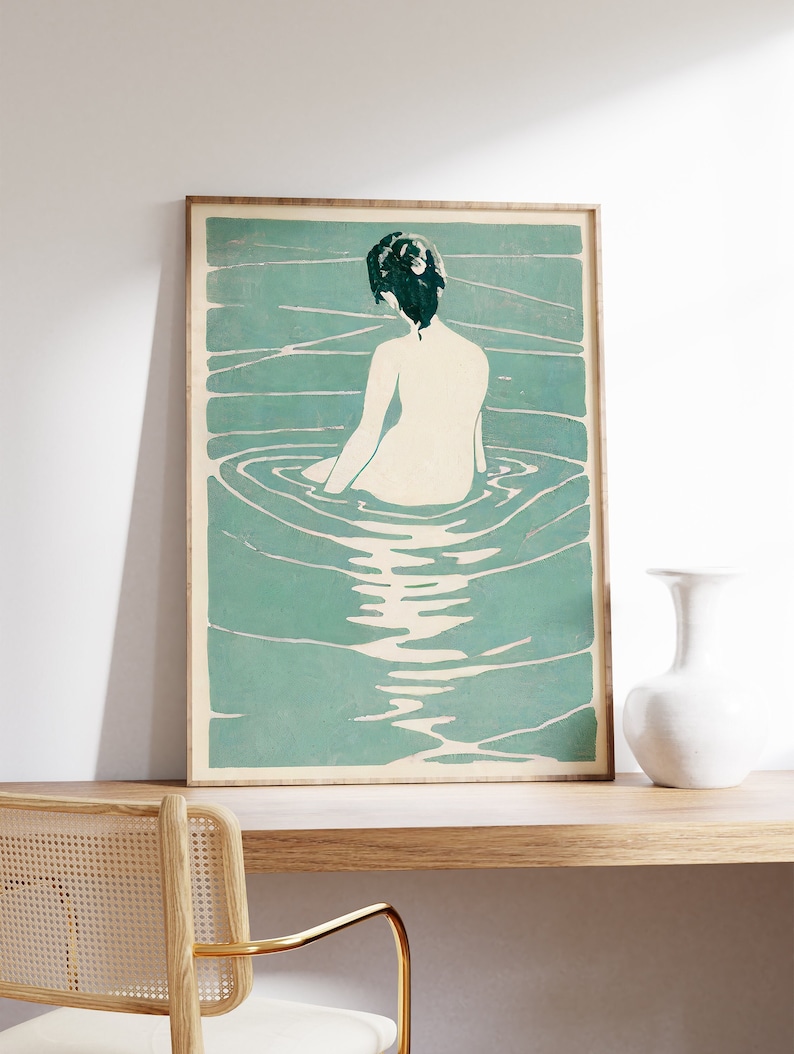 May include: A framed print of a woman sitting in a pool of water. The woman is facing away from the viewer and has dark hair. The water is a light blue-green color and has ripples around the woman.