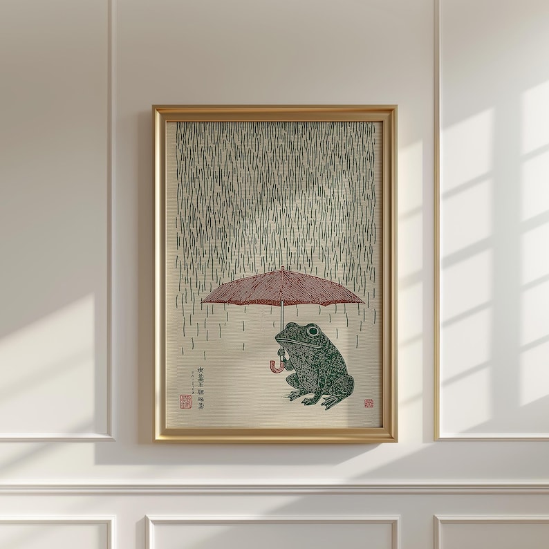 May include: Framed art print depicting a green frog sheltering under a red umbrella in the rain. The frog is illustrated in a traditional Japanese woodblock style. The artwork is set against a beige background with vertical lines, and framed in a gold-coloured frame.
