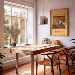 May include: A sunlit interior featuring a wooden desk with metal legs and two woven chairs. A framed artwork with Japanese text hangs on the wall. A large window offers a view of trees. A silver pendant light hangs above the desk.