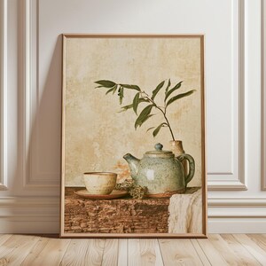 Könnte beinhalten: Ein gerahmter Kunstdruck mit einem Stillleben aus einer gesprenkelten Keramik-Teekanne und einer passenden Teetasse auf einer Untertasse, platziert auf einer rustikalen Holzoberfläche mit einem belaubten Zweig in einer Vase und weißem Stoff.