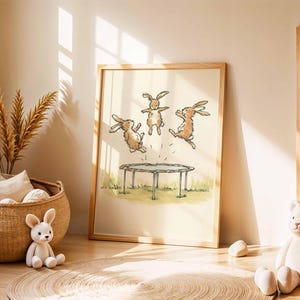 May include: A framed print featuring three cartoon rabbits jumping on a trampoline. The artwork is in a light, airy style with a wooden frame. The scene is set on a grassy area, with a neutral background. The print is displayed in a child's room.