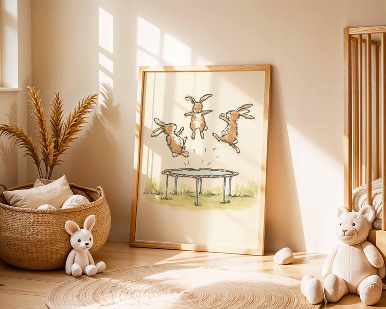 May include: A framed print featuring three cartoon rabbits jumping on a trampoline. The artwork is in a light, airy style with a wooden frame. The scene is set on a grassy area, with a neutral background. The print is displayed in a child's room.