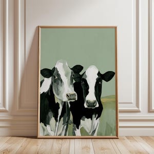 May include: A framed art print depicting two cows with black and white patterns, set against a muted green backdrop. The artwork has a rustic, farmhouse aesthetic, ideal for adding a touch of country charm to any room. The cows are painted in a semi-realistic style.