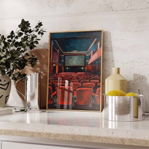 May include: A framed print of a movie theater interior with red seats and a large screen. The print is on a white countertop with a silver pitcher and a bowl of lemons.