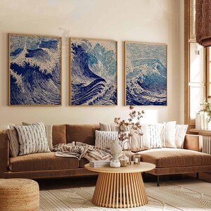 May include: Three framed art prints depicting stylized ocean waves in shades of blue and white. The artwork hangs above a brown sofa with decorative pillows. A round wooden coffee table and a woven rug complete the living room setting.