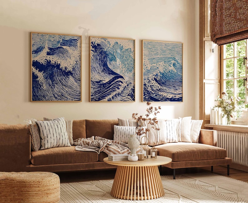 May include: Three framed art prints depicting stylized ocean waves in shades of blue and white. The artwork hangs above a brown sofa with decorative pillows. A round wooden coffee table and a woven rug complete the living room setting.