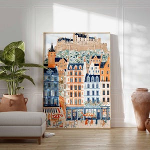 May include: A framed watercolour print depicting a European townscape with colourful buildings, a castle, and a street scene with people and umbrellas. The artwork is in a gold frame and is leaning against a white wall.