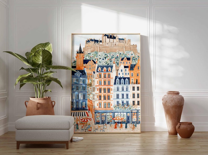 May include: A framed watercolour print depicting a European townscape with colourful buildings, a castle, and a street scene with people and umbrellas. The artwork is in a gold frame and is leaning against a white wall.