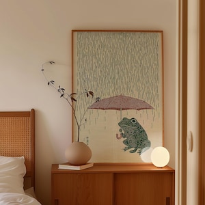May include: A framed print depicting a green frog sheltering under a red umbrella in the rain. The background has a pattern of vertical lines. The print is displayed on a wooden cabinet, alongside a vase and a spherical lamp.