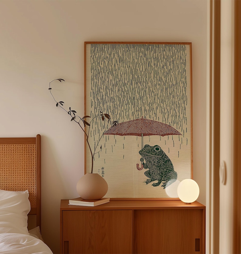 May include: A framed print depicting a green frog sheltering under a red umbrella in the rain. The background has a pattern of vertical lines. The print is displayed on a wooden cabinet, alongside a vase and a spherical lamp.
