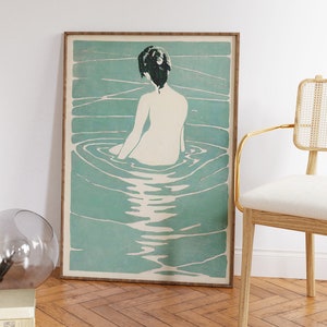 May include: A framed print of a woman sitting in a pool of water. The woman is facing away from the viewer and has dark hair. The water is a light blue-green color and has ripples around the woman.