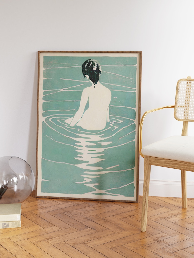 May include: A framed print of a woman sitting in a pool of water. The woman is facing away from the viewer and has dark hair. The water is a light blue-green color and has ripples around the woman.