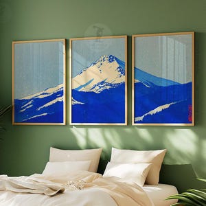 K&ouml;nnte beinhalten: Drei gerahmte Kunstdrucke, die eine Berglandschaft in Blau- und Wei&szlig;t&ouml;nen zeigen. Das Kunstwerk hat eine Vintage-&Auml;sthetik mit einem goldenen Rahmen. Die Drucke werden &uuml;ber einem Bett mit wei&szlig;er Bettw&auml;sche ausgestellt.