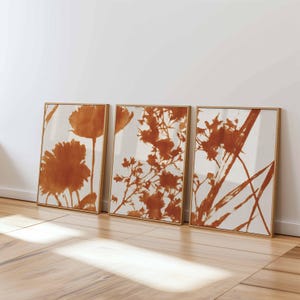 May include: Three framed art prints featuring botanical designs in shades of rust and white. The prints showcase stylized floral and plant silhouettes, each in a rectangular wooden frame. The artwork leans against a white wall on a wooden floor.