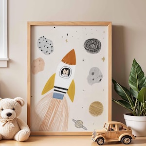 May include: A framed art print featuring a whimsical space scene. A white rocket with yellow and orange accents has a cat in the window. The print includes planets, stars, and asteroids. A teddy bear, a wooden car, and a potted plant are in the foreground.