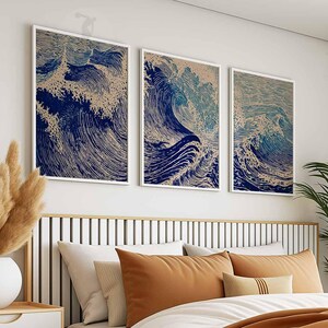 May include: Three framed art prints depicting stylized ocean waves in shades of blue and beige. The artwork is displayed above a bed with orange and white bedding. The prints are in white frames.