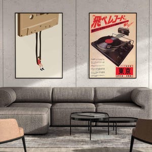 Op de afbeelding: Twee ingelijste posters op een witte muur boven een grijze hoekbank. De poster links toont een cartoonillustratie van een persoon die aan een cassetteband hangt. De poster rechts toont een vintage advertentie voor een platenspeler met de tekst "飛ベルレコード" in rood en zwart.