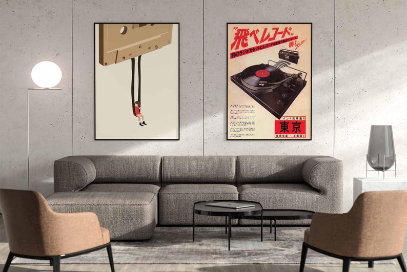 Op de afbeelding: Twee ingelijste posters op een witte muur boven een grijze hoekbank. De poster links toont een cartoonillustratie van een persoon die aan een cassetteband hangt. De poster rechts toont een vintage advertentie voor een platenspeler met de tekst "飛ベルレコード" in rood en zwart.