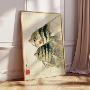 K&ouml;nnte beinhalten: Gerahmter Kunstdruck mit zwei stilisierten Fischen mit schwarz-wei&szlig;en Streifen und roten Akzenten. Das Kunstwerk hat eine Vintage-&Auml;sthetik mit beigem Hintergrund und Holzrahmen. Der Druck lehnt an einer Wand und einem Fenster.