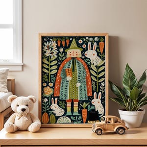 May include: A framed art print featuring a whimsical illustration of a figure in a green coat and hat, holding a book, surrounded by rabbits, carrots, and flowers. The artwork has a dark blue background and is set in a light wood frame. A teddy bear and toy car are in the foreground.