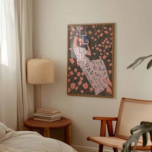 K&ouml;nnte beinhalten: Ein gerahmter Kunstdruck zeigt eine Person in einem Blumenkleid, die ein Buch in einem Feld mit rosa Blumen liest. Das Kunstwerk befindet sich in einem Holzrahmen und h&auml;ngt an einer wei&szlig;en Wand. Eine beige Lampe und ein kleiner Beistelltisch sind im Vordergrund.