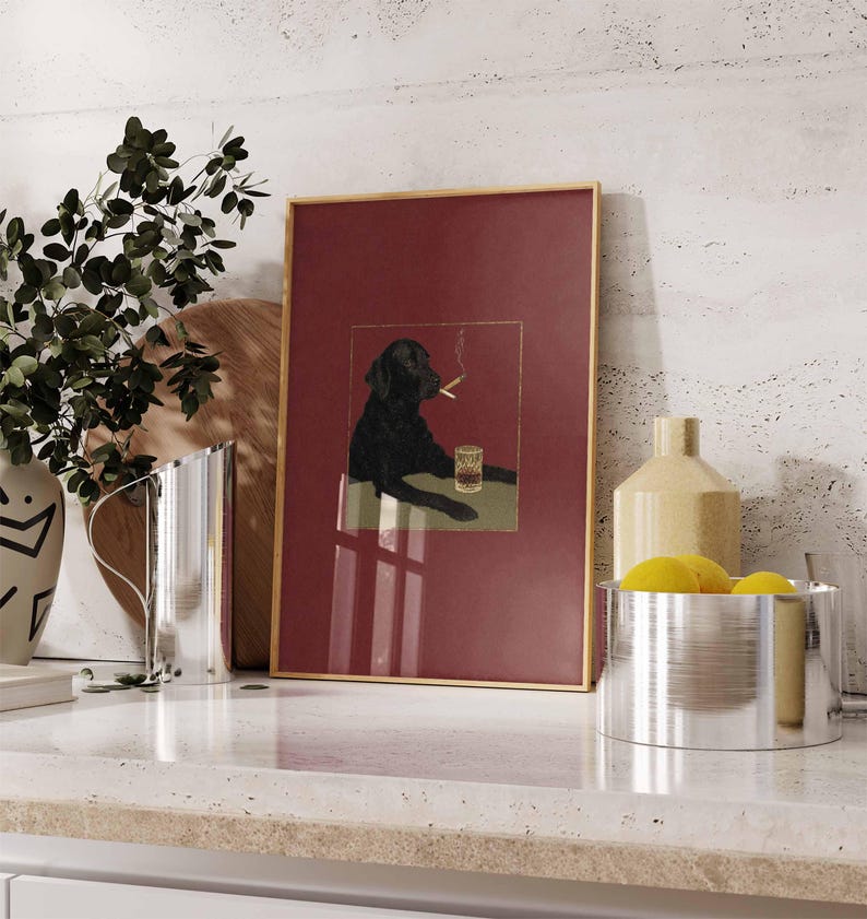 Puede incluir: L&aacute;mina art&iacute;stica enmarcada que representa a un Labrador negro fumando un cigarrillo y un vaso de whisky. La obra de arte tiene un fondo rojo y un marco dorado. La imagen se muestra sobre una encimera blanca.