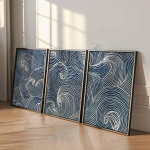 May include: Three framed abstract art pieces. Each features a swirling pattern of white lines on a dark blue background. The frames are black with a silver inner border. The artwork leans against a white wall on a wooden floor.