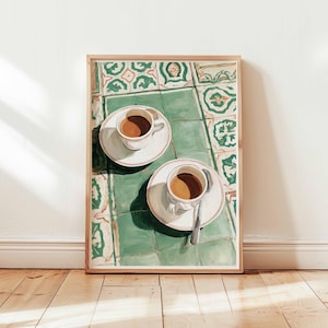 May include: A framed print depicts two cups of coffee on saucers, with a spoon on one. The cups sit on a green and white tiled surface. The artwork is in a wooden frame, leaning against a white wall and wooden floor.