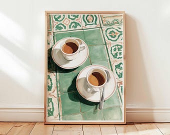 May include: A framed print depicts two cups of coffee on saucers, with a spoon on one. The cups sit on a green and white tiled surface. The artwork is in a wooden frame, leaning against a white wall and wooden floor.