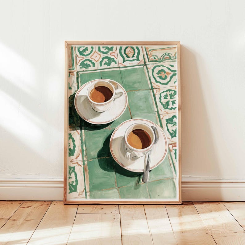 May include: A framed print depicts two cups of coffee on saucers, with a spoon on one. The cups sit on a green and white tiled surface. The artwork is in a wooden frame, leaning against a white wall and wooden floor.
