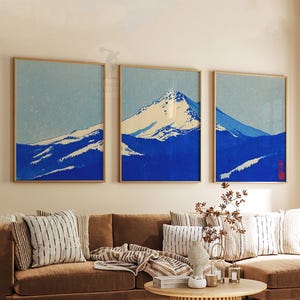 K&ouml;nnte beinhalten: Drei gerahmte Kunstdrucke, die eine Bergkette in Blau- und Wei&szlig;t&ouml;nen darstellen. Das Kunstwerk hat eine japanische &Auml;sthetik, mit einem hellblauen Himmel und einem schneebedeckten Berggipfel. Die Rahmen sind hellholzfarben.