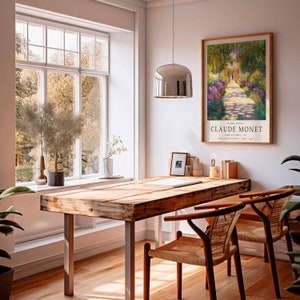 May include: A wooden table with a metal frame sits in a room with a large window and a framed print of a Claude Monet painting titled "Garden at Giverny" hanging on the wall. The print features a path lined with flowers leading to a house in the distance.