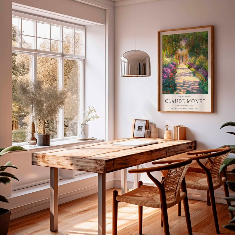 May include: A wooden table with a metal frame sits in a room with a large window and a framed print of a Claude Monet painting titled "Garden at Giverny" hanging on the wall. The print features a path lined with flowers leading to a house in the distance.