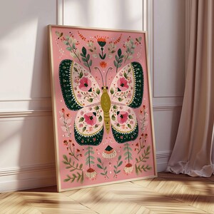 May include: A framed art print featuring a stylized butterfly with intricate floral patterns. The butterfly has pink, green, and yellow details against a pink background. The artwork is surrounded by additional floral elements and is displayed in a light wooden frame.