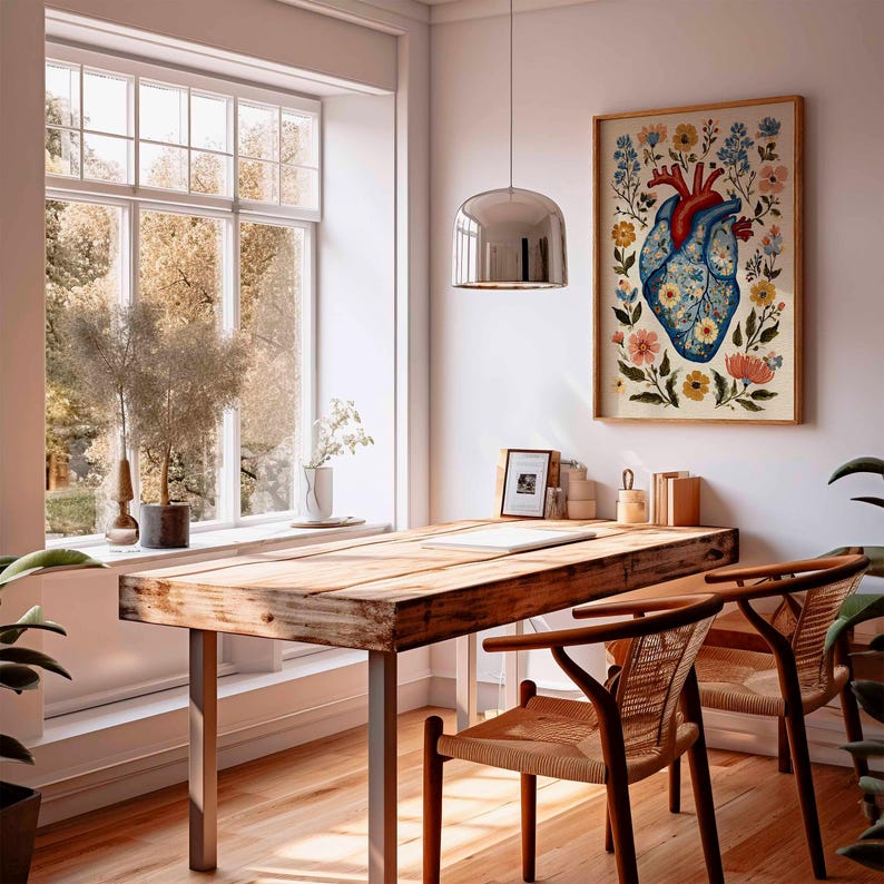 May include: A framed art print featuring a stylized anatomical heart adorned with colorful flowers. The artwork hangs above a wooden desk with two woven chairs. The room has a large window and a modern pendant light.