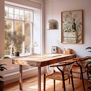 May include: A rustic wooden desk with metal legs and two woven chairs sits in a bright room. A framed artwork of olives and a bottle hangs on the wall. A large window offers a view of trees. A chrome pendant light hangs above the desk.