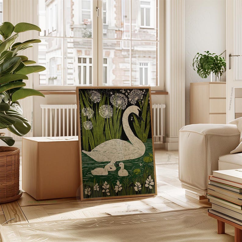 May include: Framed art print featuring a white swan with two cygnets in a pond, surrounded by green reeds and dandelions. The artwork has a black background and is displayed in a light brown wooden frame. The art is in a room with a large window.
