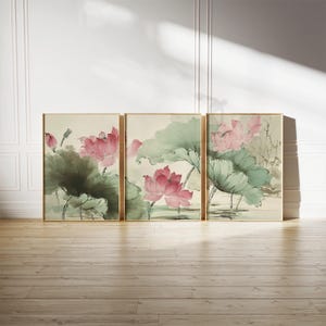 May include: Three framed art prints featuring watercolor paintings of pink lotus flowers and large green leaves. The artwork has a traditional Asian style, with gold frames and a neutral background. The prints are displayed on a wooden floor.