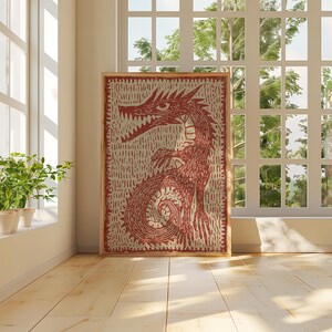 May include: A framed artwork featuring a red dragon illustration. The dragon is depicted with sharp teeth and a long tail, set against a textured, off-white background. The artwork is displayed in a room with natural light.