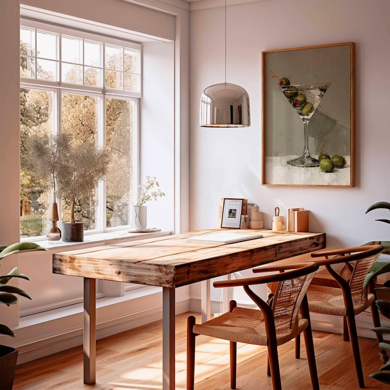 May include: A bright, airy interior scene featuring a wooden desk with metal legs, two woven chairs, and a framed painting of a martini with olives. A large window offers a view of trees, and a silver pendant light hangs above the desk.