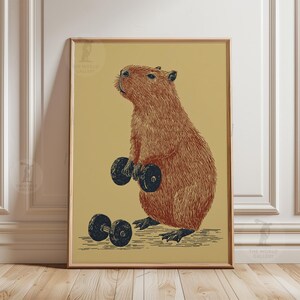 Könnte beinhalten: Ein gerahmter Druck, der ein braunes Capybara zeigt, das zwei schwarze Kurzhanteln hebt. Das Capybara ist in einem detaillierten, Vintage-Stil vor einem beigen Hintergrund dargestellt. Der Druck ist in einem goldenen Rahmen.