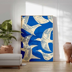 May include: A framed art print featuring a flock of birds in flight against a vibrant blue background. The birds are depicted in a cream color with detailed linework. The artwork is displayed in a room with a plant, ottoman, and decorative vases.