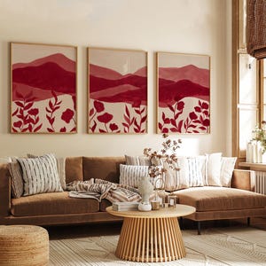 May include: Three framed wall art pieces with a mountain and floral design in shades of red and white. The artwork hangs above a brown sofa with decorative pillows. A round wooden coffee table and a woven rug complete the living room setting.