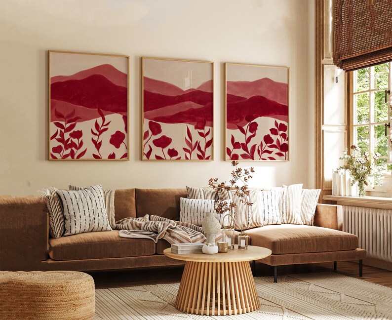 May include: Three framed wall art pieces with a mountain and floral design in shades of red and white. The artwork hangs above a brown sofa with decorative pillows. A round wooden coffee table and a woven rug complete the living room setting.