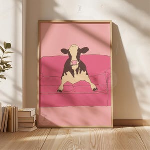 May include: Framed art print showing a cow relaxing on a pink sofa. The cow is illustrated in black and white, set against a pink backdrop and sofa. The artwork is presented in a wooden frame, offering a contemporary and playful feel.
