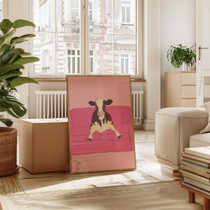 May include: A framed art print depicting a cow relaxing on a pink sofa. The cow is black and white, set against a pink backdrop. The artwork is displayed in a bright room with a large window.