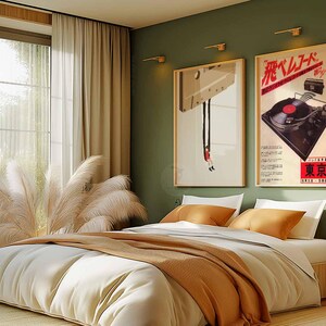 Op de afbeelding: Een slaapkamerinterieur met een bed, een raam en twee ingelijste posters aan de muur. Beide posters zijn vintage advertenties voor platenspelers. De poster aan de linkerkant toont een platenspeler die aan het plafond hangt met een persoon die eronder op de grond zit. De poster aan de rechterkant toont een platenspeler met de tekst "飛 レ コ ー ド" in het Japans. Het bed is opgemaakt met witte lakens en een bruine deken. Er liggen twee kussens op het bed, een wit en een bruin.